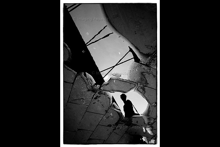 A human figure is silhouetted in a puddle on worn roadway in Lucca, Italy.